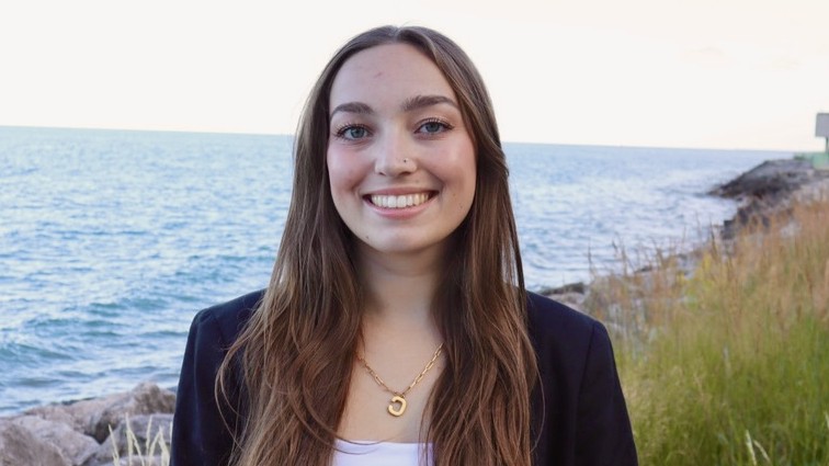 Cally Navarro stands smiling in front of Lake Michigan. She is wearing a black blazer, white shirt, and a gold necklace. Cally Navarro stands smiling in front of Lake Michigan. She is wearing a black blazer, white shirt, and a gold necklace.